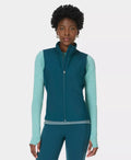 Woman wearing a teal vest over a light blue long-sleeve shirt on a white background