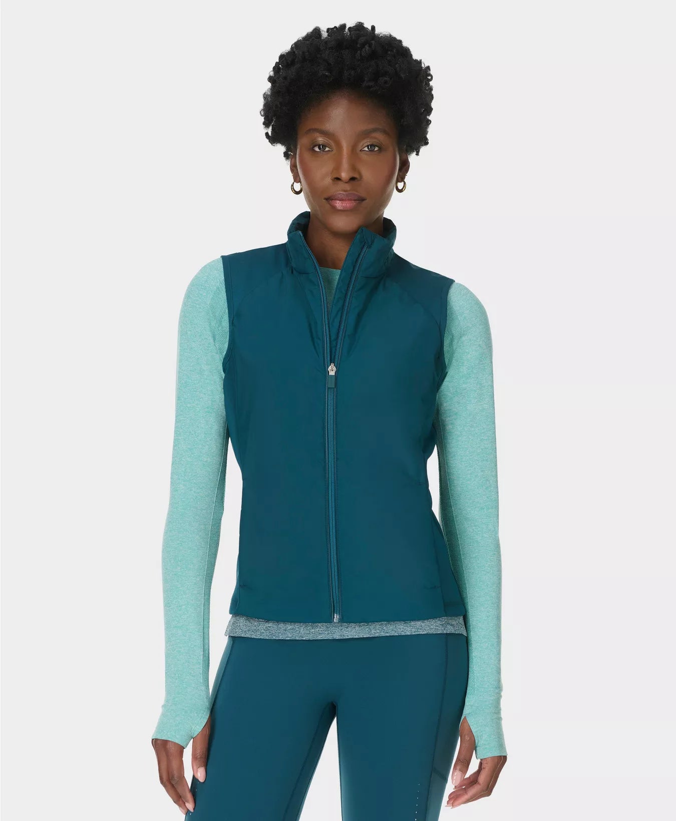 Woman wearing a teal vest over a light blue long-sleeve shirt on a white background