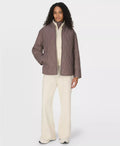 Woman wearing a taupe  brown quilted jacket and white pants on a white background
