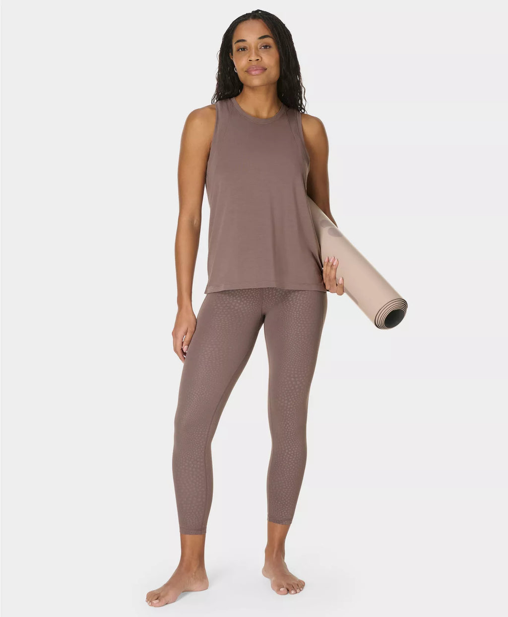 Woman in brown sleeveless top and leggings holding a yoga mat on a white background