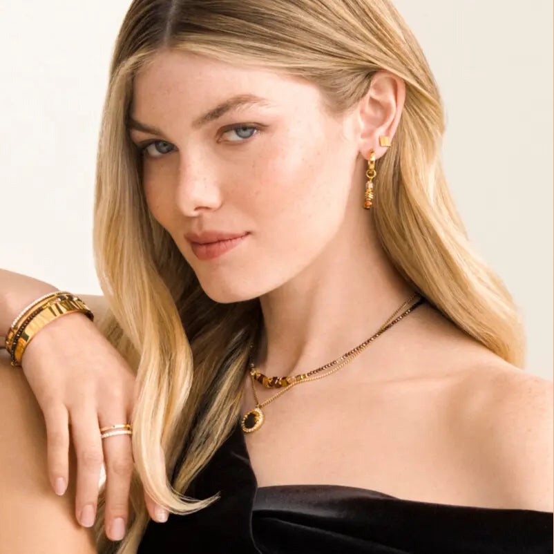 A woman with blonde hair and blue eyes wearing gold jewelry, including a necklace, earrings, bracelets, and rings, against a light background.